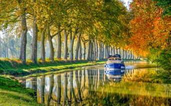 Weißes Boot auf dem Canal du Midi, gesäumt von Platanen in Herbstfarben. Die Bäume und das Boot spiegeln sich im ruhigen Wasser.