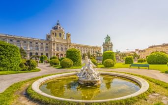 Circular fountain with white sculpture in manicured park. Kunsthistorisches Museum Vienna with dome and equestrian statue visible behind.