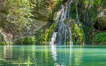Wasserfall, der über moosbewachsene Felsen in ein türkisfarbenes Becken im Petaloudes-Tal, Rhodos, fällt, umgeben von üppiger Vegetation.