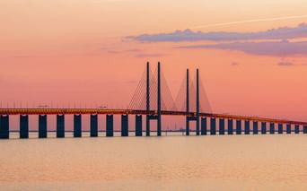 Die Öresundbrücke erstreckt sich über ruhiges Wasser unter einem rosa-orangefarbenen Sonnenuntergangshimmel. Die Schrägseilbrücke mit hohen Pylonen.