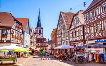 Fußgängerzone mit traditionellen Fachwerkhäusern, Kirchturm, Straßencafés und Radfahrern. Bunte Gebäude säumen Kopfsteinpflasterstraße.