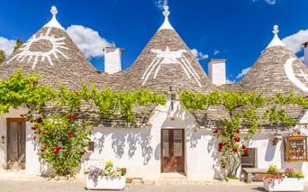 Weiße Trulli-Häuser mit konischen Steindächern und bemalten Symbolen in Alberobello, geschmückt mit Kletterpflanzen und bunten Blumen.