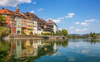 Historische bunte Fachwerkhäuser der Laufenburger Altstadt spiegeln sich im ruhigen Flusswasser. Kirchturm ragt über den Gebäuden auf.