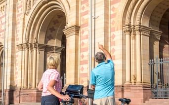 Zwei Radfahrer mit Tourenrädern und blauen Taschen stehen vor der gestreiften romanischen Fassade des Speyerer Doms mit verzierten Portalen.