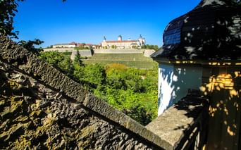 Blick auf die Festung Marienberg in Würzburg von Steinbefestigungen, mit Weinbergen am Hang und blauem Himmel. Historischer Turm im Vordergrund.