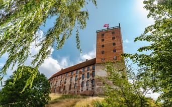Koldinghus Schloss in Kolding mit rotem Backsteinturm und dänischer Flagge, eingerahmt von grünen Bäumen unter blauem Himmel mit weißen Wolken.