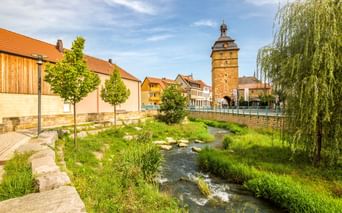 Bach fließt durch Bad Staffelstein Innenstadt mit historischem Steinturm, bunten Häusern und orangefarbenen Ziegeldächern unter blauem Himmel.