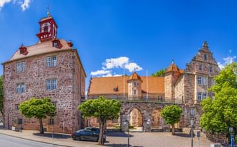 Landgrafenschloss Eschwege mit roten Sandsteingebäuden, Glockenturm und orangen Ziegeldächern. Bäume säumen den Kopfsteinpflasterhof unter blauem Himmel.