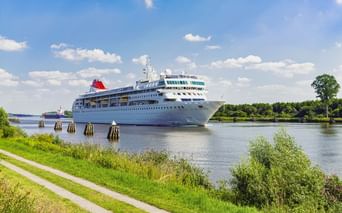 Weißes Kreuzfahrtschiff mit rotem Schornstein auf dem Nord-Ostsee-Kanal. Grüne Ufer mit Radweg und Holzpfosten säumen die Wasserstraße unter blauem Himmel.