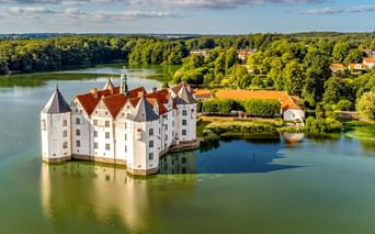 Schloss Glücksburg, ein weißes Renaissance-Wasserschloss mit roten Dächern, umgeben von einem See. Grüne Wälder und blauer Himmel im Hintergrund.