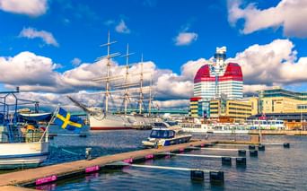 Hafen von Göteborg mit weißem Segelschiff, modernen Gebäuden mit rot-weißer Fassade, Holzstegen und schwedischer Flagge unter blauem Himmel.