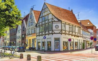 Traditionelles Fachwerkhaus an einer Ecke mit orangefarbenem Ziegeldach in Glückstadt. Die weiße Fassade zeigt dunkle Holzbalken und Geschäfte im Erdgeschoss.