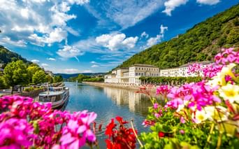 Colorful flowers frame the Lahn River in Bad Ems with historic white buildings along the waterfront, green hills, and a boat moored at the shore.