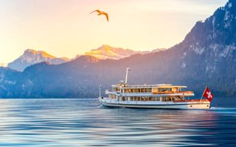 Weißes Passagierschiff mit Schweizer Flagge auf dem Genfer See bei Sonnenaufgang, mit Bergen im Hintergrund und einem Vogel am Himmel.