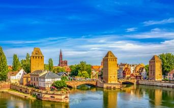 Ponts Couverts in Straßburg mit drei mittelalterlichen Türmen entlang eines Kanals, Brücke und historischen Gebäuden unter blauem Himmel.