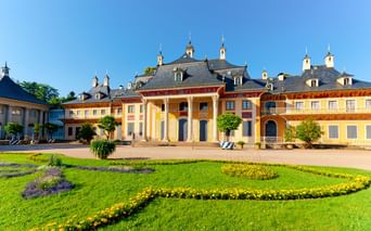 Schloss Pillnitz mit gelber Fassade und weißen Säulen, umgeben von gepflegten Rasenflächen mit bunten Blumenbeeten unter blauem Himmel.
