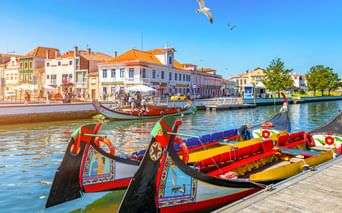 Traditionelle Moliceiro-Boote mit bunten Verzierungen im Kanal von Aveiro. Historische Gebäude mit orangen Dächern säumen das Ufer unter blauem Himmel.