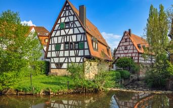 Traditionelle bayerische Fachwerkhäuser mit orangen Ziegeldächern spiegeln sich in einem ruhigen Fluss in Hersbruck, umgeben von grünen Bäumen.