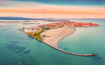 Luftaufnahme der Halbinsel Grado in der türkisfarbenen Adria mit Sandstrand, rotgedeckten Gebäuden und buntem Sonnenuntergangshimmel.