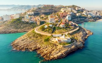 Luftaufnahme von Cullera, Spanien, zeigt eine felsige Halbinsel mit weißen Gebäuden, kurvenreichen Küstenstraßen und türkisfarbenem Mittelmeer.