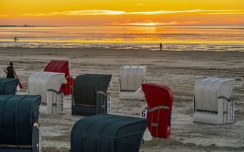 Traditionelle Strandkörbe am Strand in Bensersiel bei Sonnenuntergang. Goldener Himmel spiegelt sich im Nordseewasser mit Spaziergängern in der Ferne.