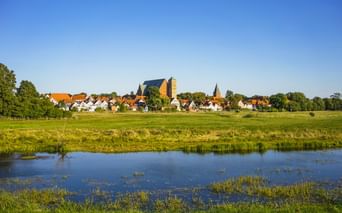 Blick auf Verden an der Aller mit historischer Kirche und Turm über weißen Häusern mit roten Dächern. Grüne Wiesen und Fluss im Vordergrund.