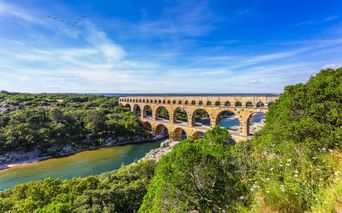Der Pont du Gard, eine dreistöckige römische Aquäduktbrücke über einem grünen Flusstal, umgeben von dichter mediterraner Vegetation unter blauem Himmel.