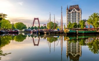 Historischer Hafen in Rotterdam mit vertäuten Segelschiffen, die sich im ruhigen Wasser spiegeln. Rote Willemsbrücke und Witte Huis sichtbar.