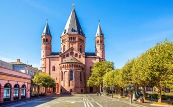 Mainzer Dom mit drei markanten Türmen und roter Sandsteinfassade auf einem sonnigen Platz mit grünen Bäumen und blauem Himmel.