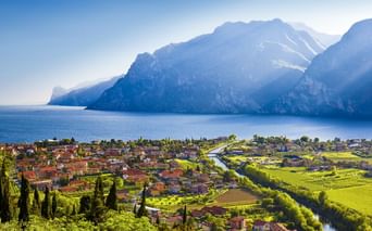 Dorf Pietramurata im Sarcatal mit rotgedeckten Häusern, grünen Feldern und Gardasee, umgeben von dramatischen Berggipfeln unter blauem Himmel.