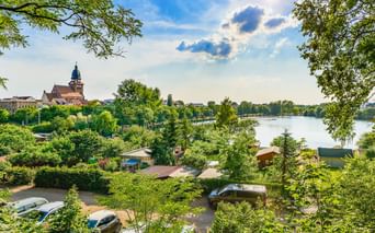 Blick durch grüne Bäume auf eine Stadt am See mit Kirchturm und historischen Gebäuden. Ein Campingplatz ist im Vordergrund sichtbar.