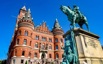 Rotes Backsteinrathaus Helsingborg mit verzierten Türmen, mit einer bronzenen Reiterstatue im Vordergrund vor klarem blauen Himmel.