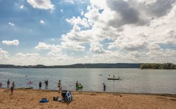 Sandstrand am Brombachsee mit schwimmenden und spielenden Menschen im Wasser. Blauer Himmel mit weißen Wolken über dem See und bewaldeter Uferlinie.