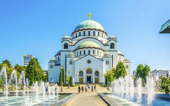 Dom des Heiligen Sava in Belgrad mit weißer Fassade, grünen Kupferkuppeln und goldenen Kreuzen. Springbrunnen säumen den Weg zum Eingang.