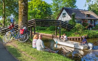 Zwei Radfahrer sitzen an einem Kanal in Giethoorn und beobachten ein Boot unter einer Holzbrücke. Fahrräder stehen in der Nähe, ein reetgedecktes Haus ist sichtbar.