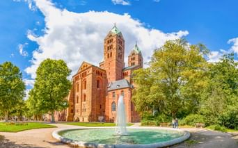 Speyerer Dom mit roten Sandsteintürmen und romanischer Architektur, umgeben von grünen Bäumen. Ein runder Brunnen mit Fontäne im Vordergrund.