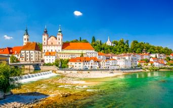 Steyrer Altstadt in Österreich mit barocken Kirchtürmen, bunten Gebäuden mit orangen Dächern entlang eines türkisfarbenen Flusses mit Wasserfall.