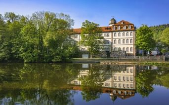 Weißes Barockschloss in Rotenburg an der Fulda mit orangefarbenem Dach und Turm, spiegelt sich im ruhigen Flusswasser. Grüne Bäume rahmen das Gebäude unter blauem Himmel.