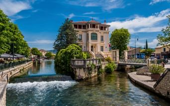 Rosa Villa mit grünen Fensterläden neben einem fließenden Kanal in L'Isle-sur-la-Sorgue, Frankreich. Ein kleiner Wasserfall und eine Brücke sind sichtbar.
