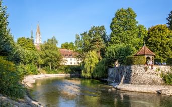 Malerischer Blick auf den Konstanzer Stadtgarten mit Fluss, Steinpavillon, üppigen Bäumen und Kirchtürmen im Hintergrund bei blauem Himmel.