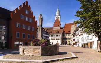 Marktplatz in Überlingen mit Steinbrunnen, rotem Giebelhaus und Kirchturm. Kopfsteinpflasterplatz mit historischen Gebäuden unter blauem Himmel.
