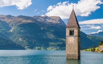 Historischer Kirchturm von Graun ragt aus dem Reschensee in Bayern, mit Bergen und blauem Himmel im Hintergrund entlang der Via Claudia Augusta.