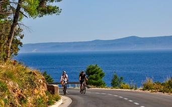 Zwei Radfahrer auf einer Küstenstraße in Dalmatien, Kroatien, mit blauer Adria und fernen Bergen unter klarem Himmel.