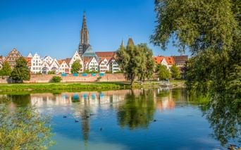 View across the Danube River to Ulm's historic old town with colorful half-timbered houses and the towering Ulm Minster spire under blue sky.