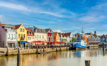 Reihe bunter historischer Häuser mit roten, gelben und weißen Fassaden entlang eines Hafenkanals. Ein blaues Boot liegt am Ufer unter blauem Himmel.
