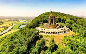 Luftaufnahme des Kaiser-Wilhelm-Denkmals auf bewaldetem Hügel in Porta Westfalica. Die Weser schlängelt sich durch grüne Felder im Tal.
