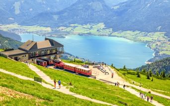 Rote Zahnradbahnen an der Schafberg Bergstation auf grüner Almwiese mit Blick auf den türkisen Wolfgangsee und Berge in Österreich.