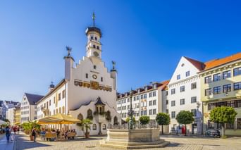 Weißes Rathaus mit Glockenturm und Brunnen auf Kopfsteinpflasterplatz in Kempten. Bunte Gebäude und Straßencafés umgeben den Platz unter blauem Himmel.