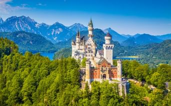 Schloss Neuschwanstein mit weißen Türmen umgeben von grünem Wald, mit Bergketten und blauen Seen im Hintergrund unter klarem Himmel.