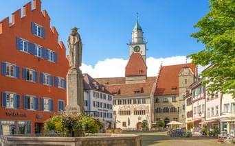 Historischer Marktplatz in Überlingen mit Steinbrunnen, Statue, bunten Gebäuden und Kirchturm unter blauem Himmel.